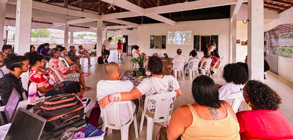 A fotografia, tirada de um ângulo posterior, mostra um grupo de aproximadamente 40 pessoas participando de uma capacitação ou palestra em um espaço amplo e coberto.