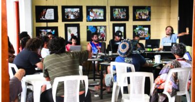 Foto de uma reunião coletiva em um espaço cultural. Um grupo diversificado de pessoas está sentado em cadeiras de plástico brancas e ao redor de mesas com computadores. Ao fundo, uma parede de tijolos amarelos exibe várias fotografias emolduradas em preto. À direita, um homem de camiseta azul gesticula enquanto fala para o grupo. O ambiente é informal e colaborativo