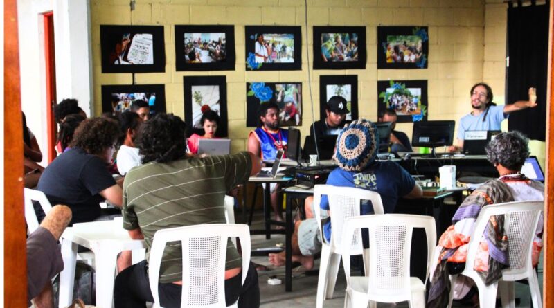 Foto de uma reunião coletiva em um espaço cultural. Um grupo diversificado de pessoas está sentado em cadeiras de plástico brancas e ao redor de mesas com computadores. Ao fundo, uma parede de tijolos amarelos exibe várias fotografias emolduradas em preto. À direita, um homem de camiseta azul gesticula enquanto fala para o grupo. O ambiente é informal e colaborativo
